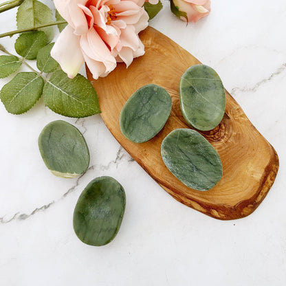 A small wooden tray holds five oval green Nephrite Jade Worry Stones with marbled patterns. One worry stone is placed near the tray on a white surface. Two light pink roses with green leaves rest beside the tray. The setting is minimalist and elegant.