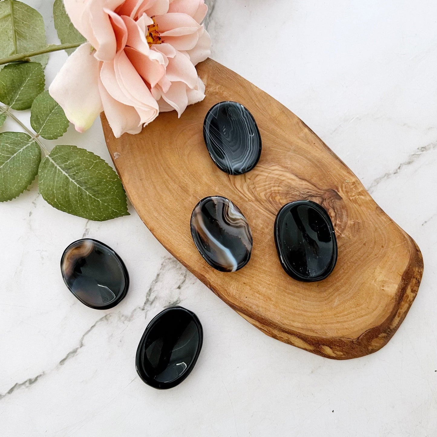 A wooden tray holds five polished oval stones, renowned as Black Onyx Worry Stones for their protective power, with various dark patterns. They're nestled next to a peach-colored rose and green leaves, all resting on a marble surface.