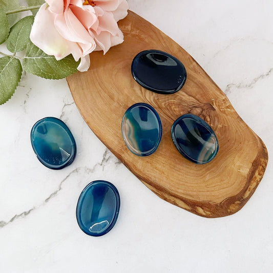 A wooden oval tray holds five polished blue and green gemstones, each with unique shades and patterns. A Blue Onyx Worry Stone stands out, offering calming influence and metaphysical benefits. A sprig of greenery with a light pink flower lies at the top left corner of the tray, all set on a white marble surface.