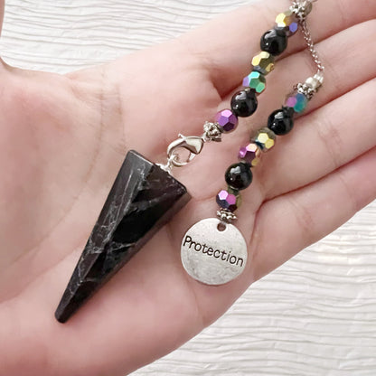 A Black Tourmaline Pendulum with Protection Charm rests on top of a black book with gold and white lettering. The divination tool, known for its grounding and protective properties, is positioned on a white textured surface.