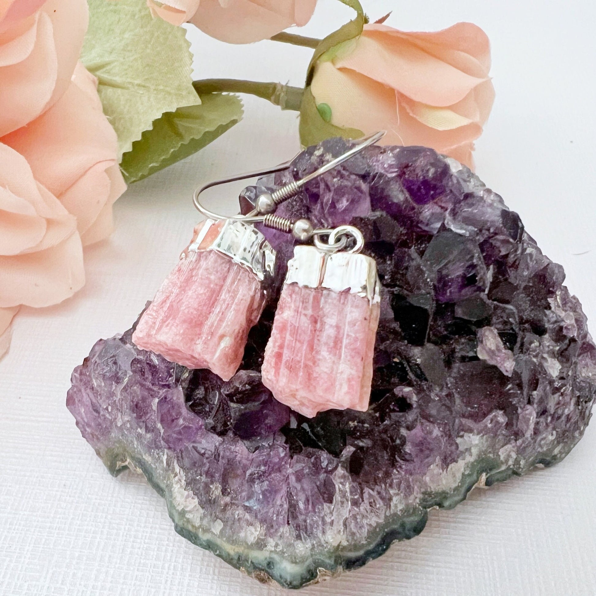 A pair of Raw Pink Tourmaline Point Dangle Earrings with silver hooks is displayed on a purple amethyst crystal cluster. In the background, there are soft pink rose flowers with green leaves, all set on a white surface. They are designed to enhance emotional balance while adding a touch of elegance.