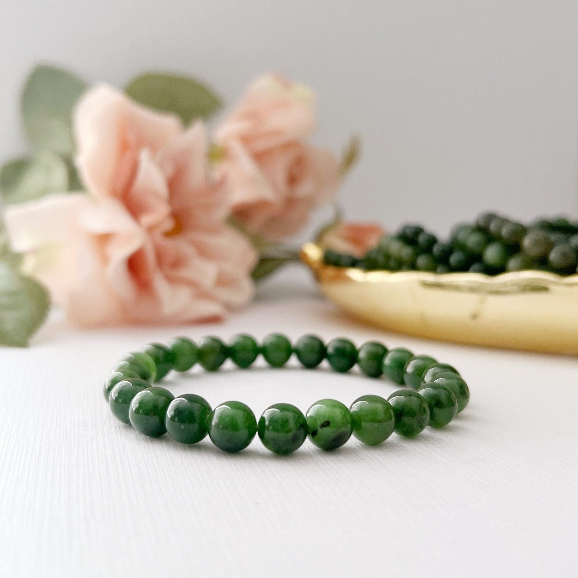 A 6-8mm Nephrite Jade Beaded Bracelet is displayed on a white surface with a backdrop of pink flowers and a gold decorative dish filled with green beads. The gemstone accessory and objects are arranged to create a serene and elegant composition, embodying harmony, balance, and abundance.