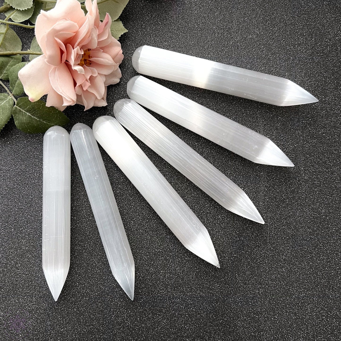 Six 5-6" Long Selenite Wands of varying lengths are arranged on a dark surface. Nearby, a pink rose with green leaves adds a touch of floral beauty to the display. Perfectly crafted for energy work, the wands have a smooth, reflective surface and are pointed at both ends.