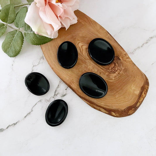 A wooden tray on a marble surface holds five polished black stones of different sizes. Among them, an Obsidian Worry Stone stands out as a protective gemstone believed to aid in emotional healing. A light pink rose with green leaves is placed next to the tray.

Notice that the product name "Obsidian Worry Stone" already appears correctly in the sentence.