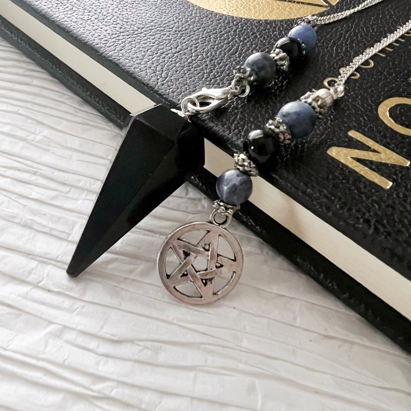 A Black Obsidian Hexagonal Pendulum with Sodalite & Pentacle Charm and a beaded chain with a silver pentagram pendant are placed on top of a closed black book. The book has gold-embossed text on the cover, partially visible, grounding energies and intuitive powers. The background features a textured white surface.