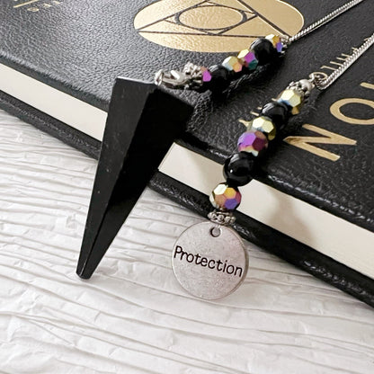 A Black Tourmaline Pendulum with Protection Charm rests on top of a black book with gold and white lettering. The divination tool, known for its grounding and protective properties, is positioned on a white textured surface.