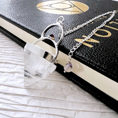 A photo of a Raw Apophyllite Pendulum with Garnet Accent & Silver Sun-Moon Embrace, lying on top of a black hardcover book with gold lettering. Perfect for spiritual divination, the pendulum is attached to a silver chain with a small charm that includes a purple bead.