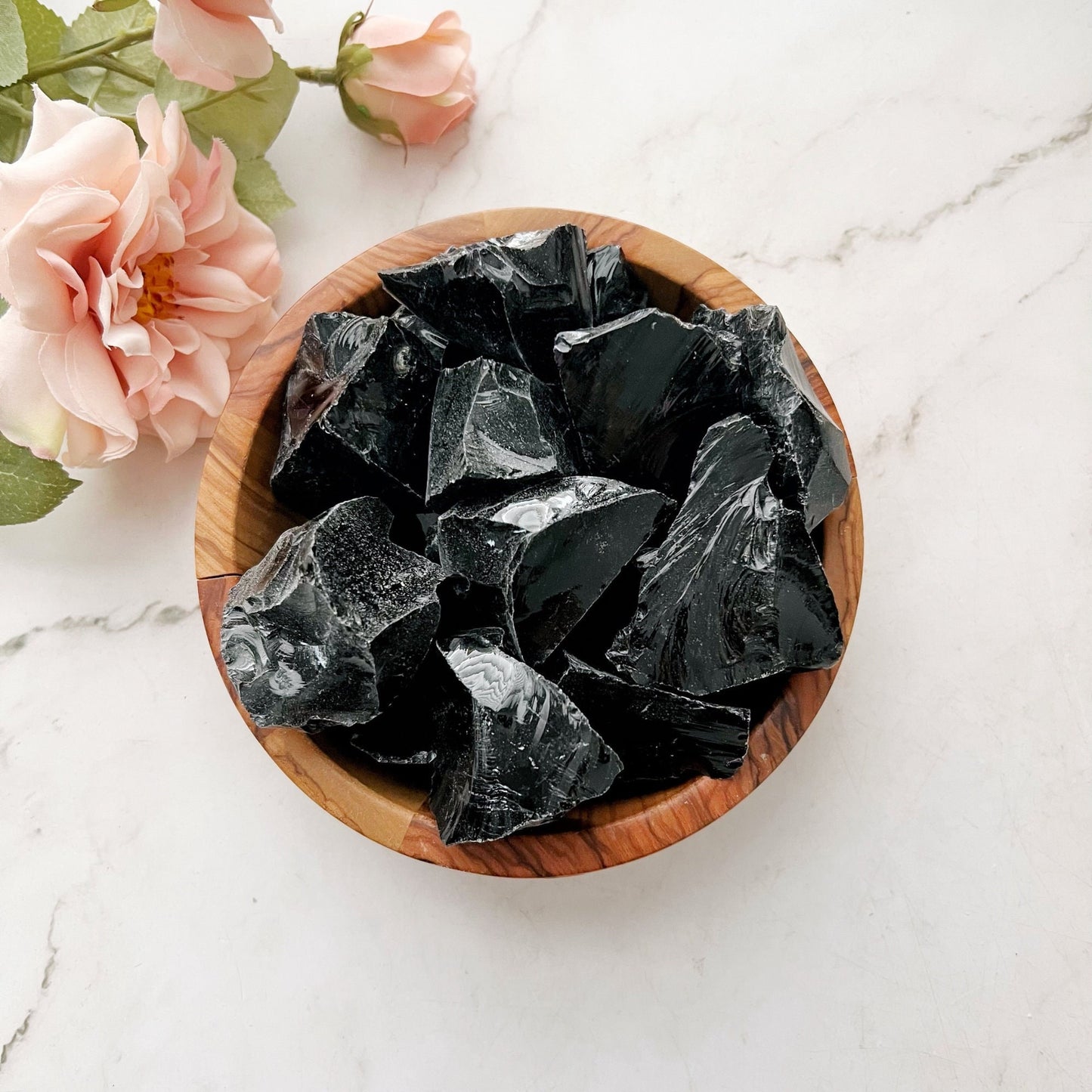 A wooden bowl filled with shiny, rough Raw Black Obsidian Stones is placed on a white marble surface. Next to the bowl, there are soft pink flowers and green leaves, enhancing the display's emotional healing ambiance and highlighting the obsidian's protective qualities.