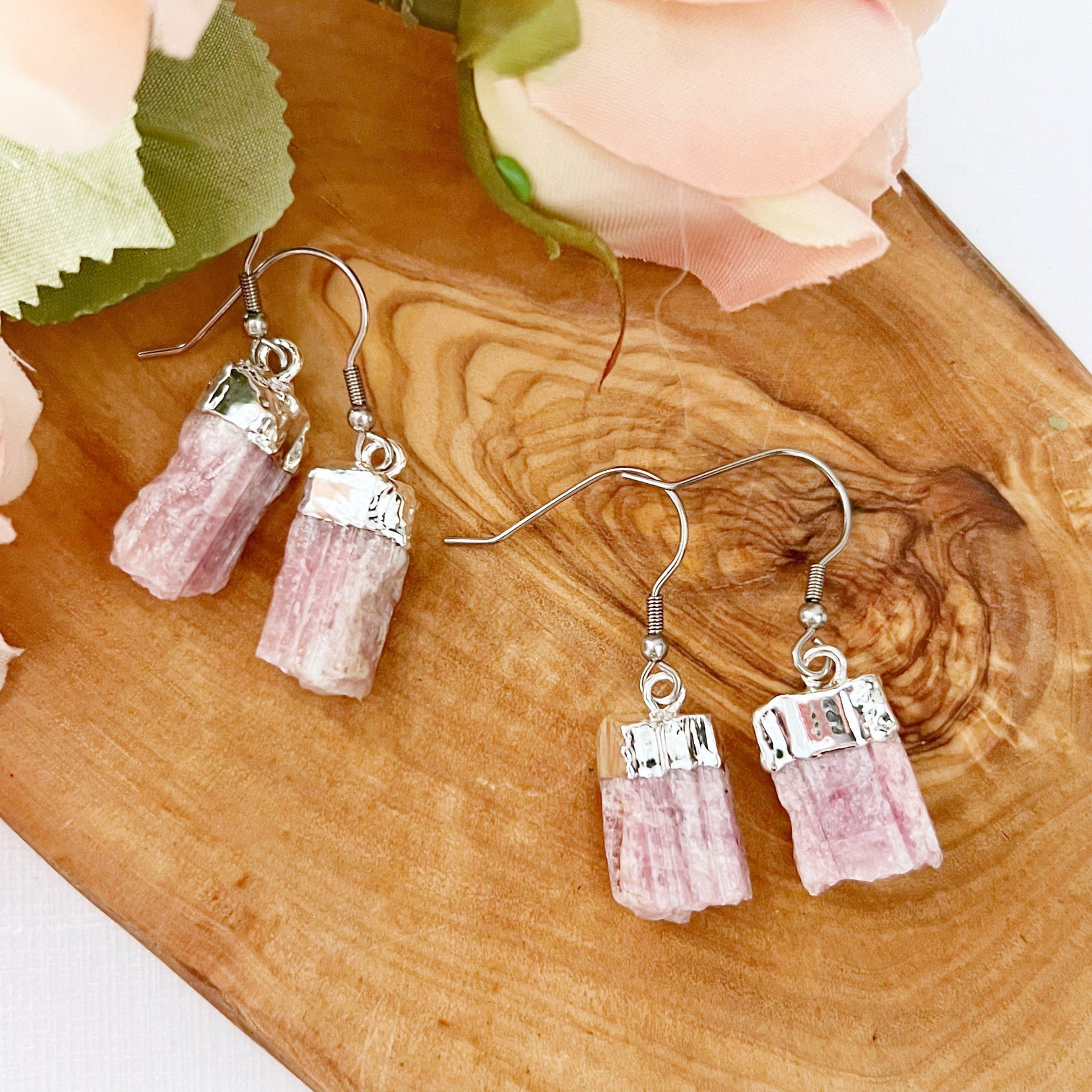 A pair of Raw Pink Tourmaline Point Dangle Earrings with silver hooks is displayed on a purple amethyst crystal cluster. In the background, there are soft pink rose flowers with green leaves, all set on a white surface. They are designed to enhance emotional balance while adding a touch of elegance.