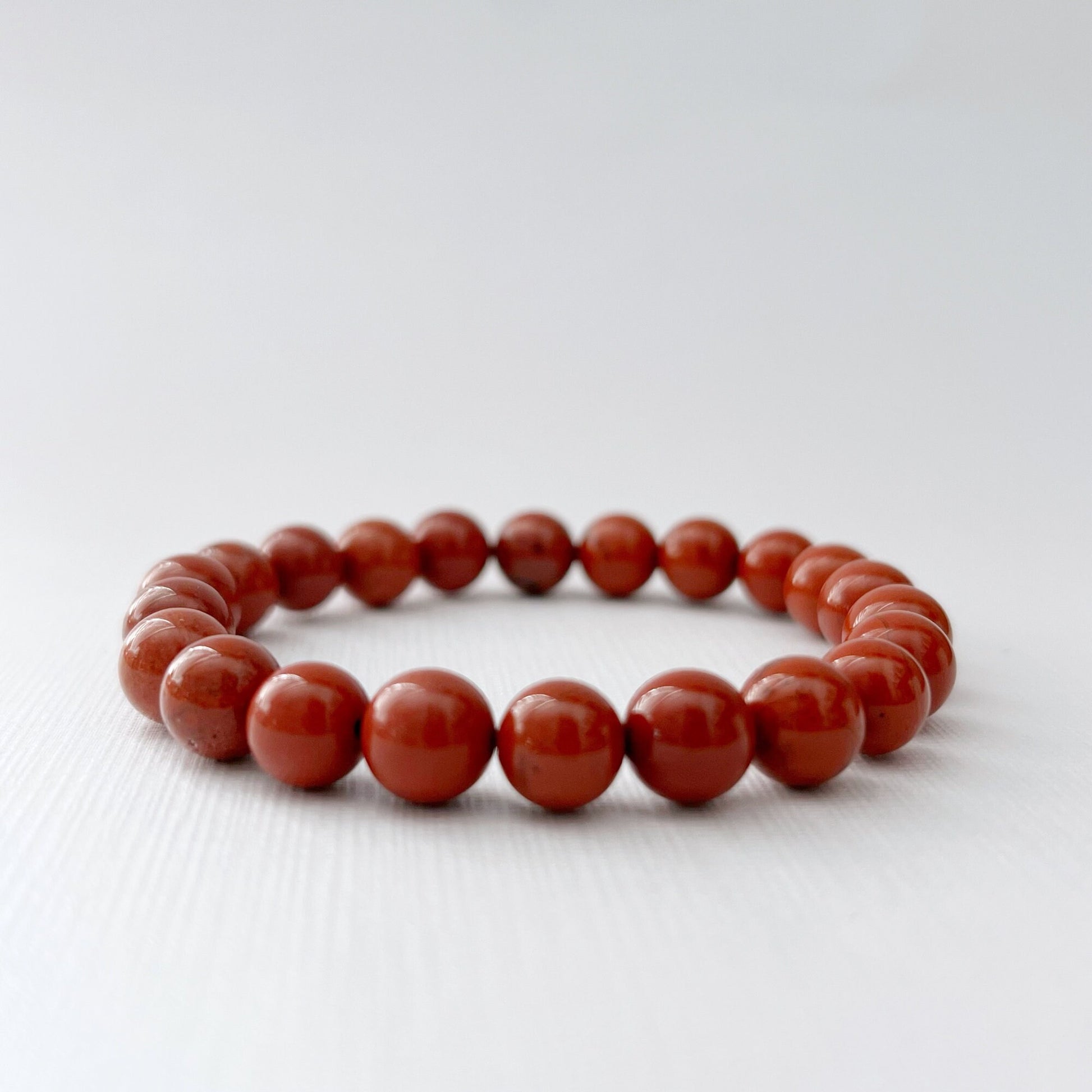 An 8mm Red Jasper Beaded Bracelet made of round, polished red beads is placed on a white surface. In the background, there are pink roses and a shallow bowl filled with more red beads. The scene is brightly lit, highlighting the grounding energy and vibrant colors of the beads and flowers.