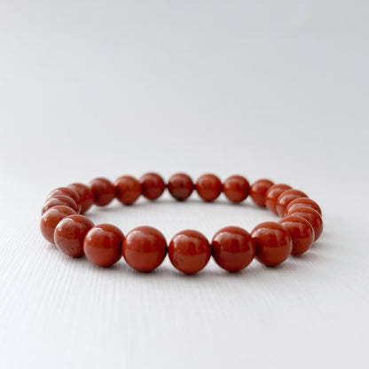 An 8mm Red Jasper Beaded Bracelet made of round, polished red beads is placed on a white surface. In the background, there are pink roses and a shallow bowl filled with more red beads. The scene is brightly lit, highlighting the grounding energy and vibrant colors of the beads and flowers.