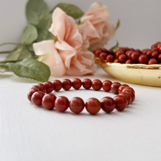 An 8mm Red Jasper Beaded Bracelet made of round, polished red beads is placed on a white surface. In the background, there are pink roses and a shallow bowl filled with more red beads. The scene is brightly lit, highlighting the grounding energy and vibrant colors of the beads and flowers.