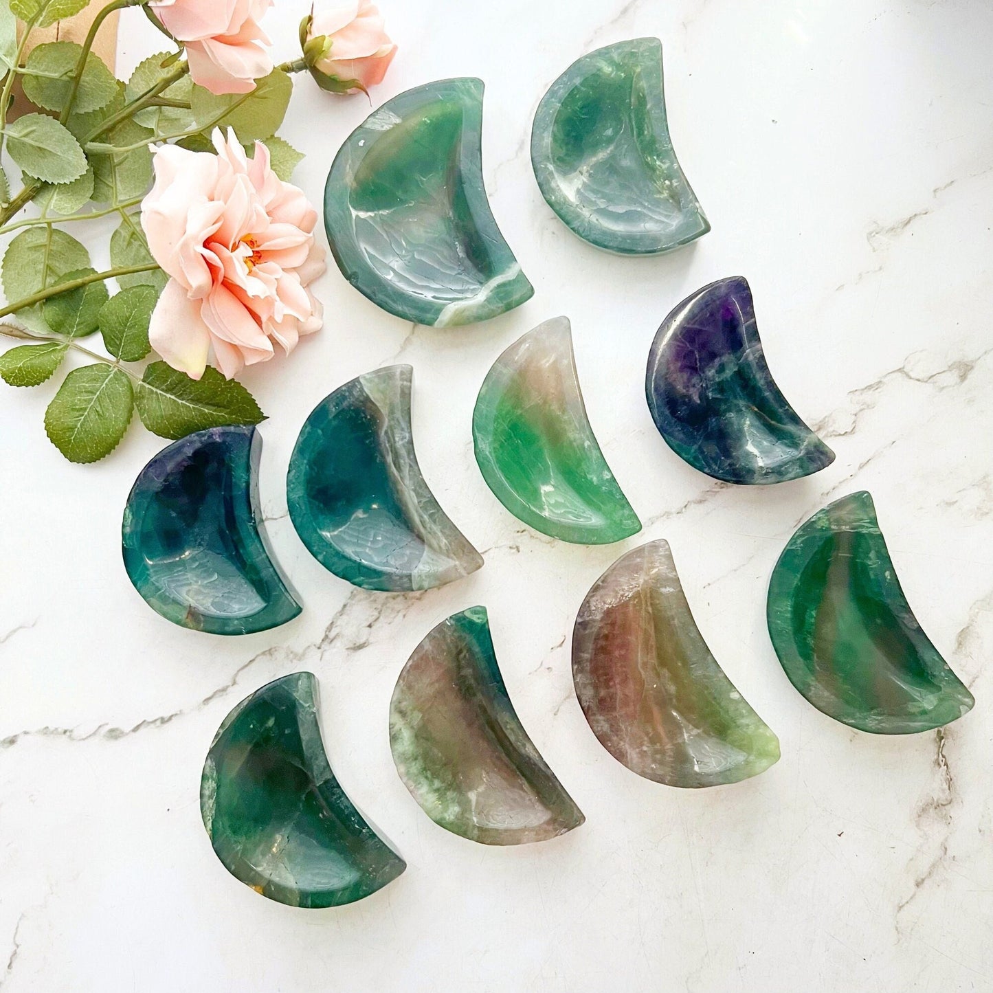 An arrangement of a 4" Rainbow Fluorite Crescent Moon Bowl in various shades of green and purple placed on a marble surface. To the top left, pink flowers with green leaves add a natural touch to the scene, enhancing its spiritual energy and metaphysical properties.