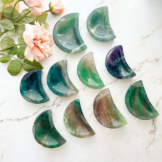 An arrangement of a 4" Rainbow Fluorite Crescent Moon Bowl in various shades of green and purple placed on a marble surface. To the top left, pink flowers with green leaves add a natural touch to the scene, enhancing its spiritual energy and metaphysical properties.