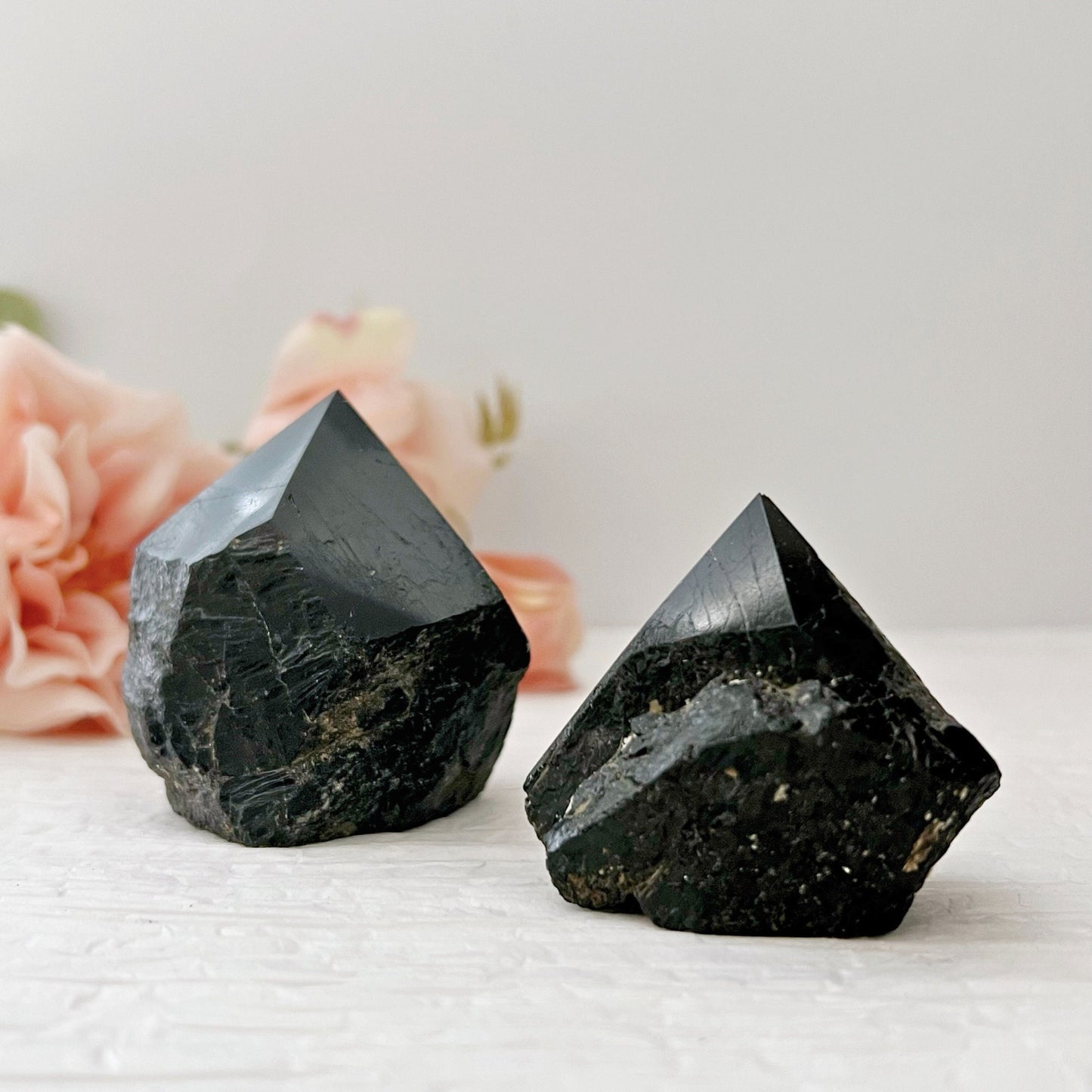 Two Black Tourmaline Top Polished Points, featuring a lustrous finish and angular facets, are displayed on a textured white surface. In the soft-focus background, pale pink flowers add a gentle contrast to the deep, dark hue of these protective properties-laden crystals.