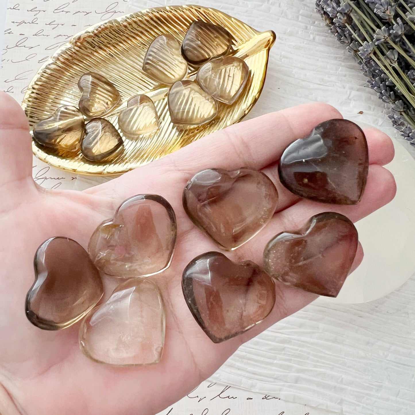 A gold dish with multiple translucent 1" Mini Smoky Quartz Crystal Heart stones sits on white paper with handwritten text. Next to the dish, a round white object adorned with three of these stones, including grounding Smoky Quartz, is arranged. Lavender flowers can be seen in the corner.