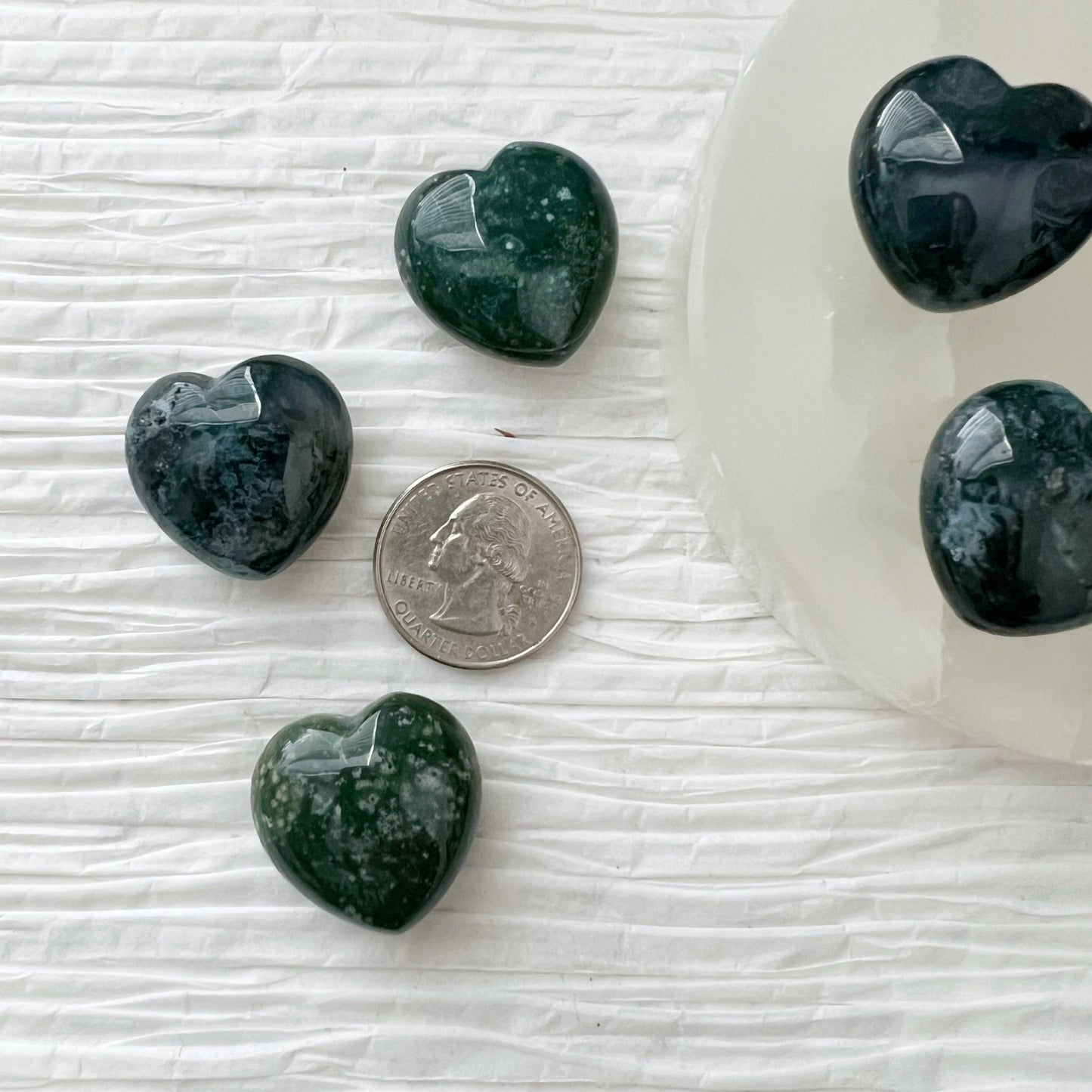 A collection of heart-shaped, 1" Mini Green Moss Agate Crystal Hearts with white speckles are displayed on a white surface. Some gemstone hearts are arranged on a round, translucent white plate, while others rest on a golden, leaf-shaped dish. Sprigs of lavender lie nearby, symbolizing new beginnings.