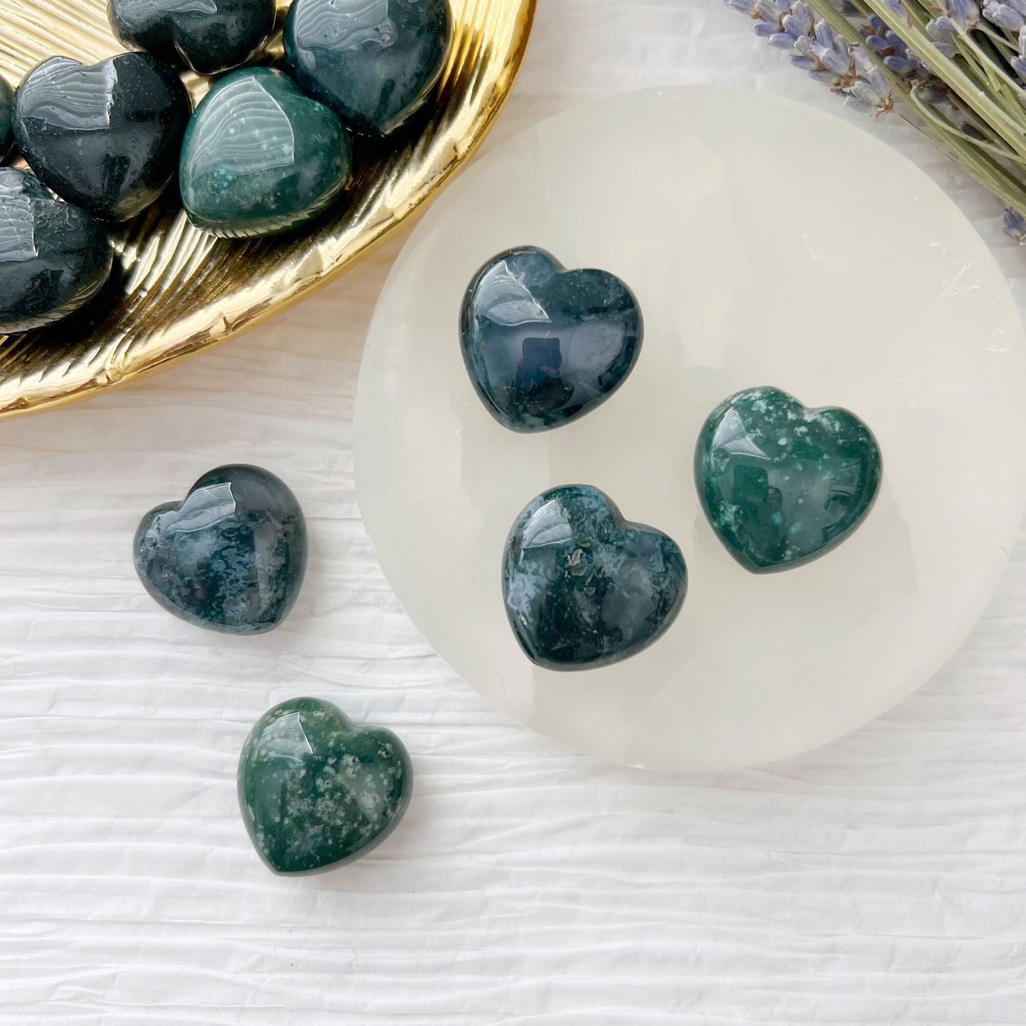 A collection of heart-shaped, 1" Mini Green Moss Agate Crystal Hearts with white speckles are displayed on a white surface. Some gemstone hearts are arranged on a round, translucent white plate, while others rest on a golden, leaf-shaped dish. Sprigs of lavender lie nearby, symbolizing new beginnings.