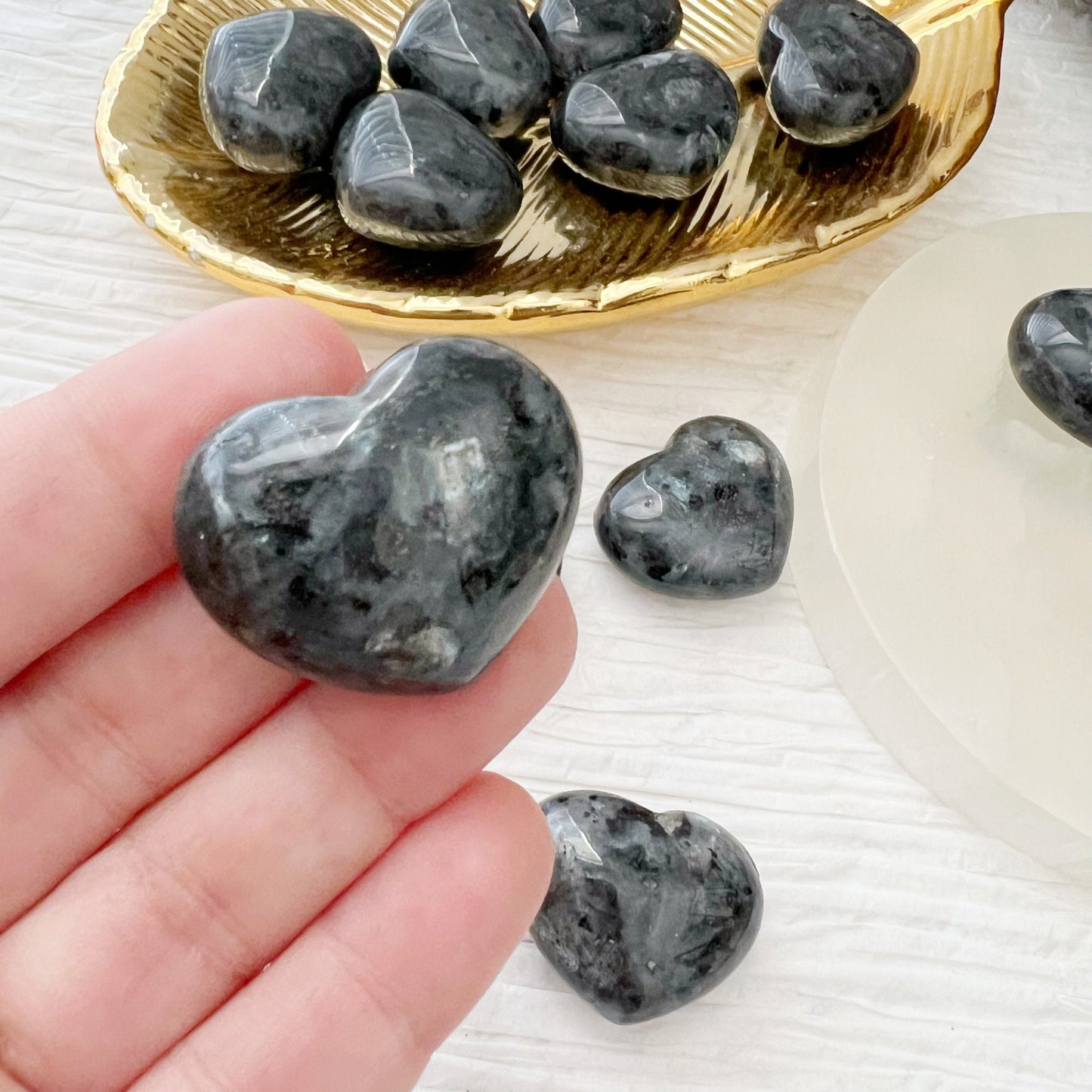 A photograph of six black 1" Mini Larvikite Crystal Hearts. Three are placed on a gold dish, and three more are on a white circular surface. Known for their grounding and protective qualities, these stones are complemented by sprigs of lavender visible at the top right corner for decoration.