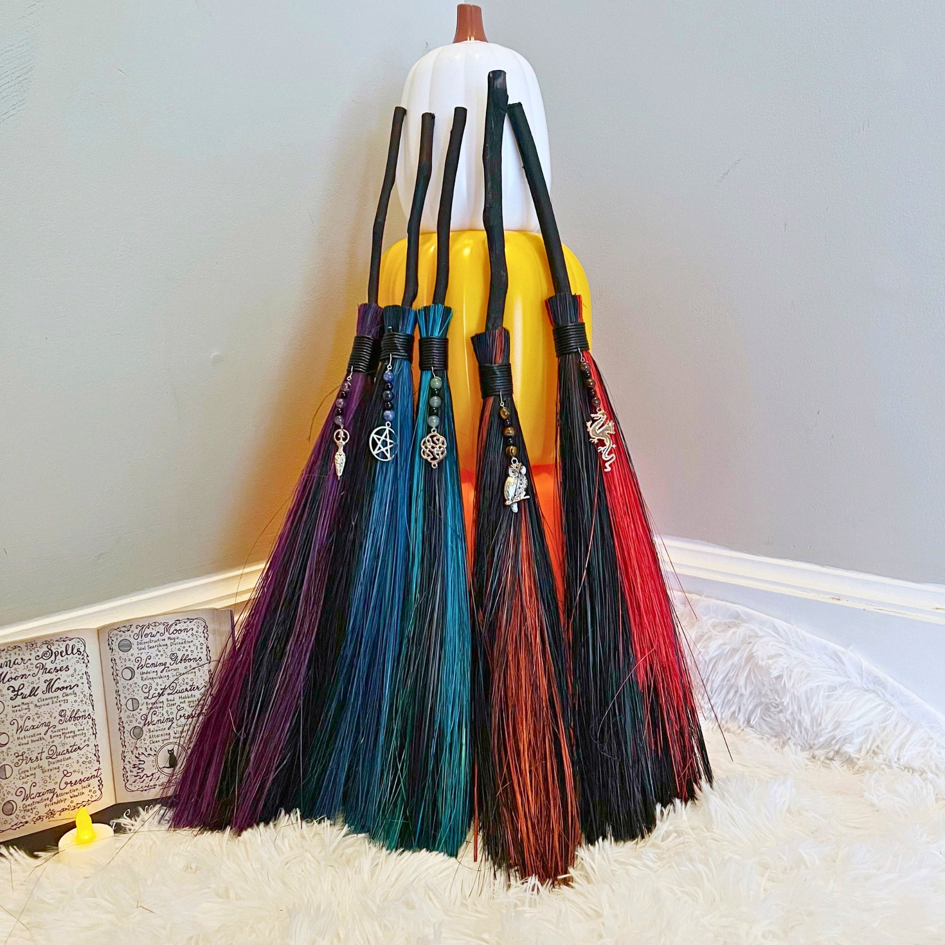 A set of six 18" Witch Besom Ritual Bamboo Broom decorations, perfect for witchy decor, are propped against a large, decorative ceramic pumpkin. Each broomstick features a different charm and vibrant bristles in shades of purple, blue, green, red, and orange. The setup is on a fluffy white rug.