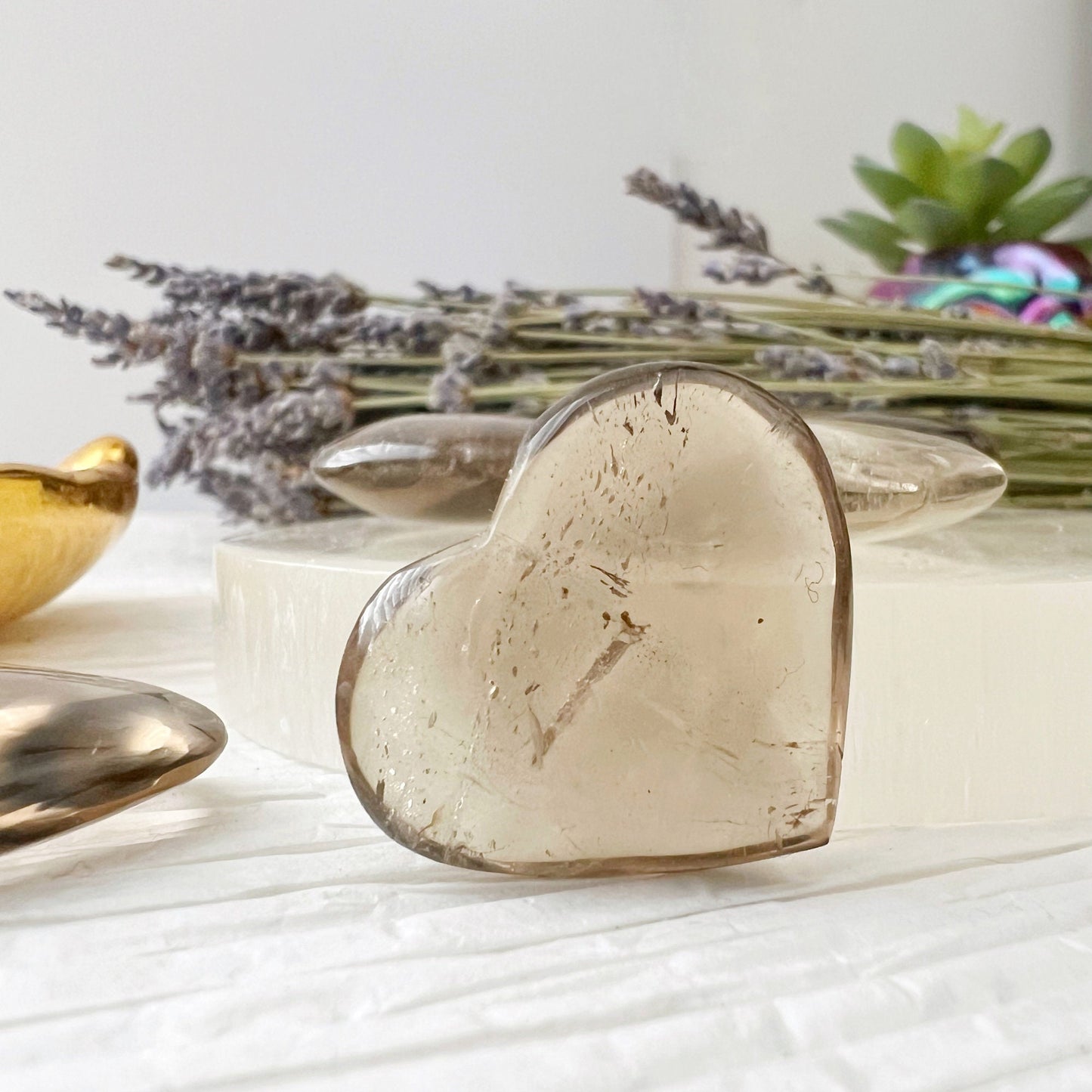 A gold dish with multiple translucent 1" Mini Smoky Quartz Crystal Heart stones sits on white paper with handwritten text. Next to the dish, a round white object adorned with three of these stones, including grounding Smoky Quartz, is arranged. Lavender flowers can be seen in the corner.