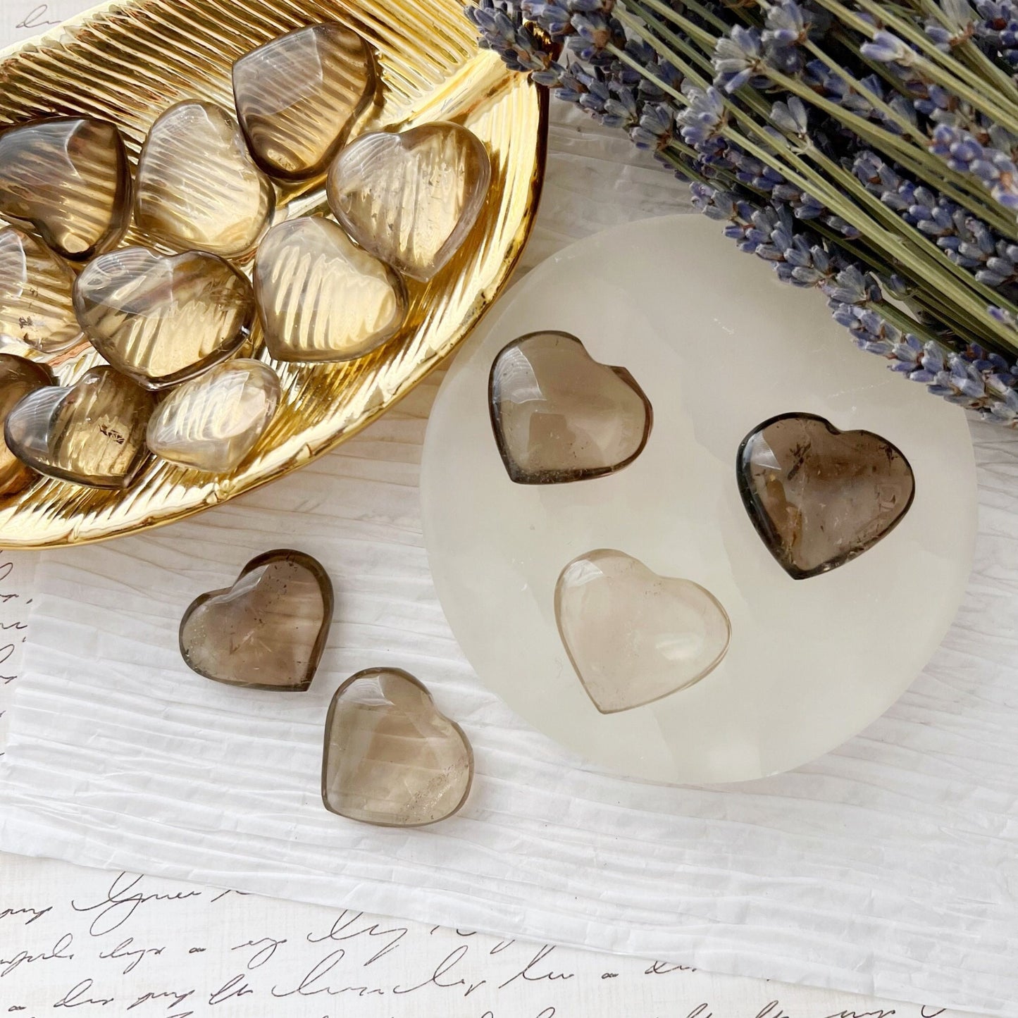 A gold dish with multiple translucent 1" Mini Smoky Quartz Crystal Heart stones sits on white paper with handwritten text. Next to the dish, a round white object adorned with three of these stones, including grounding Smoky Quartz, is arranged. Lavender flowers can be seen in the corner.