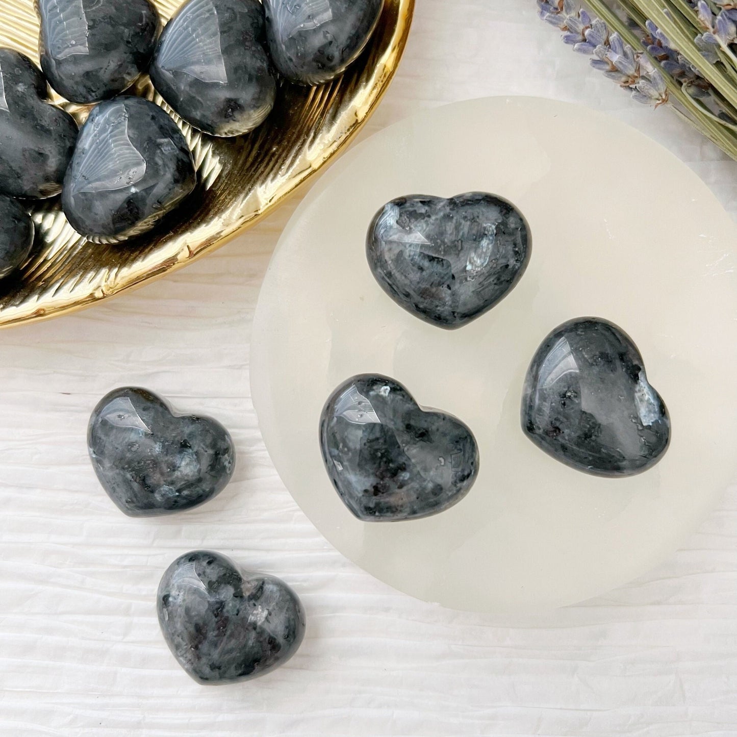 A photograph of six black 1" Mini Larvikite Crystal Hearts. Three are placed on a gold dish, and three more are on a white circular surface. Known for their grounding and protective qualities, these stones are complemented by sprigs of lavender visible at the top right corner for decoration.