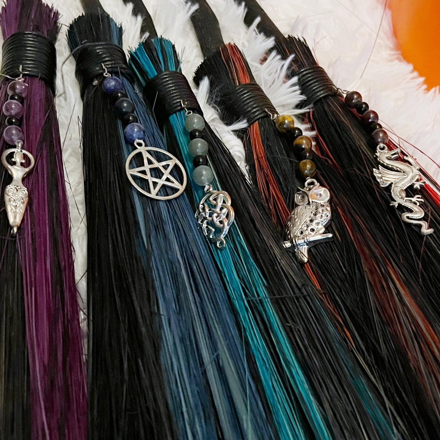 A set of six 18" Witch Besom Ritual Bamboo Broom decorations, perfect for witchy decor, are propped against a large, decorative ceramic pumpkin. Each broomstick features a different charm and vibrant bristles in shades of purple, blue, green, red, and orange. The setup is on a fluffy white rug.