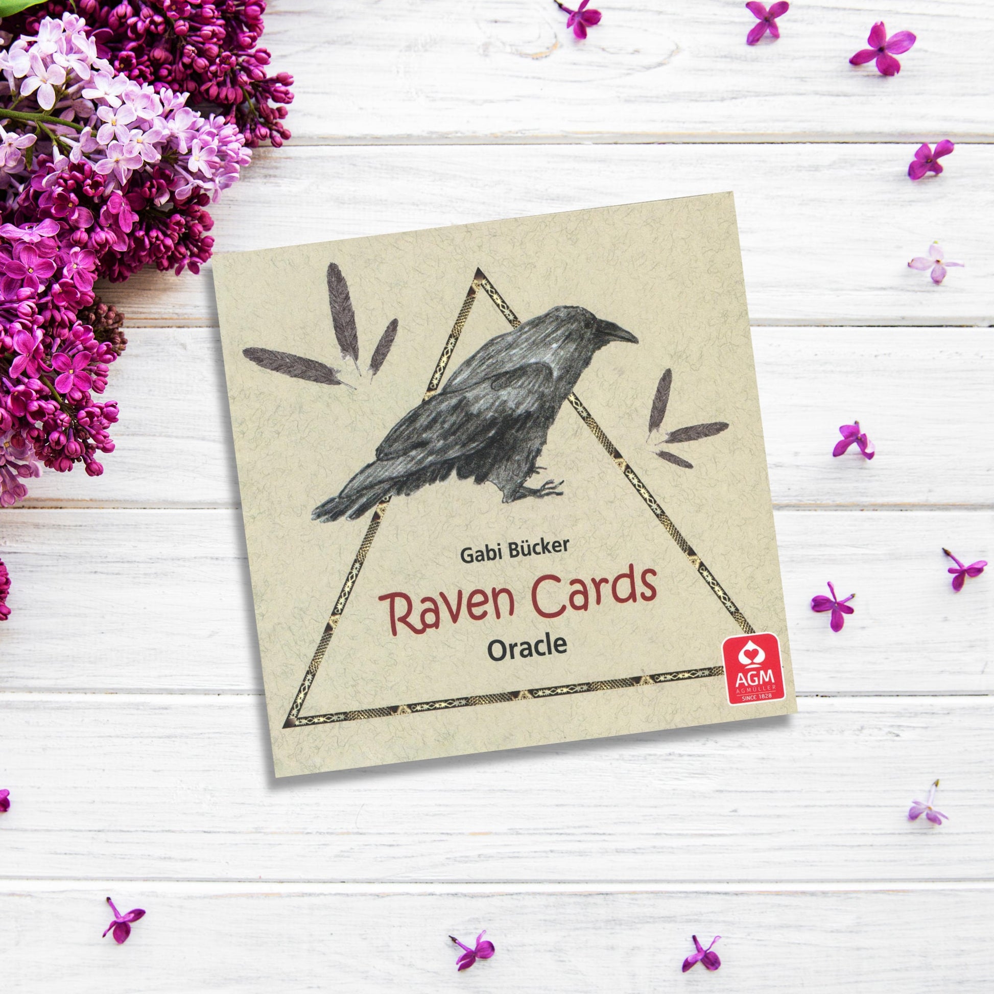 The image shows a square card with a hand-drawn illustration of a raven, labeled "Raven Cards Oracle Deck," and the AGM logo in the corner. The card is placed on a white wooden surface adorned with small purple flowers and lilac blossoms, evoking insightful messages steeped in myth and legend.